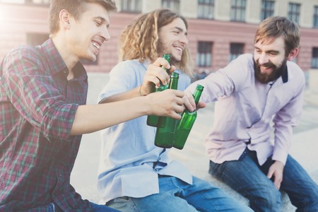 Happy friends cheers with beer outdoor. Company of young man have rest and clinking with bottles....