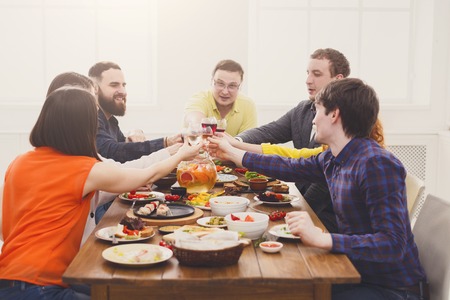 People clink glasses, saying cheers at party dinner table in cafe, restaurant or at home. Young cute friends company celebrate with organic food at wooden table indoors.の写真素材