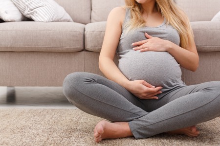 Unrecognizable pregnant woman caressing her belly at home. Young expectant blonde feeling her baby push, sitting on floor, copy space. Pregnancy, rest, life, expectation conceptの写真素材