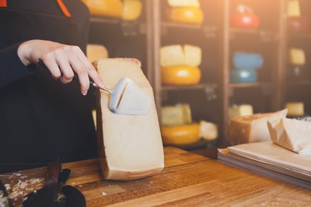 Parmesan cheese closeup. Half wheel of cheese on wooden table. Shop assistant slicing cheese with special knife, background, copy spaceの写真素材