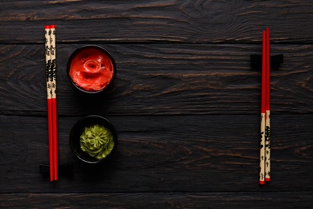 Pieces of traditional japanese tablewear. Red chopsticks with hierogliphs, small black bowls with ginger and wasabi on dark wooden table. Top view, flat lay, copy space, backgroundの写真素材