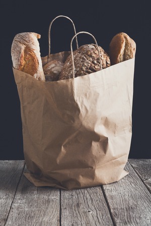 Bread assortment in paper bag on rustic wood table at black background. Shopping at bakeryの写真素材