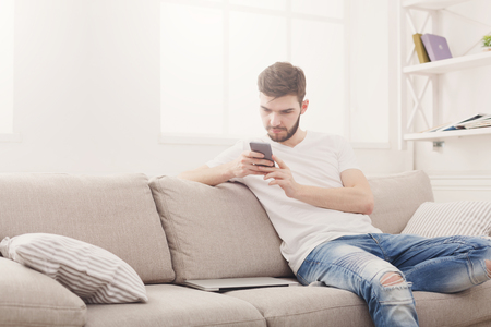 Young man at home sending messages on smartphone and laptop lying aside on beige couch in light livingroom, copy spaceの写真素材