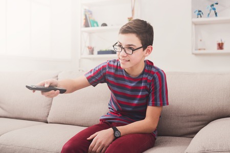 Teenager boy watching television, pointing with remote conroller while sitting on couch in living room at homeの写真素材