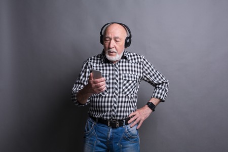 Senior man listening to music on smartphone with headphones, choosing track on mobile, gray studio background, copy spaceの写真素材