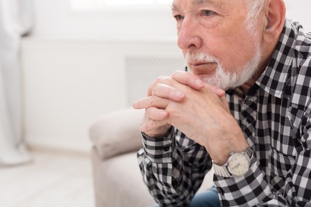 Pensive elderly man sitting on couch with thoughtful facial expression, copy space