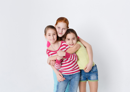 Lifestyle portrait of three smiling girls at white studio background with copy space. Friends hugging, posing on camera. Friendship, togetherness and supportの写真素材