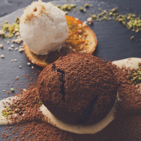 Exquisite french dessert. Chocolate fondant with creme anglaise and vanilla ice cream, sprinkled with cocoa powder, served on round slate decorated with orange citron and pistachio crumble, closeupの写真素材