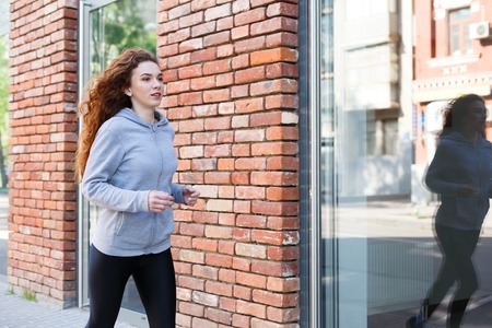 Young redhead woman jogging in city near brick building with mirror windows, copy spaceの写真素材