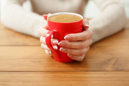 Woman holding a cup of hot drink while sitting at wooden table, shallow depth of field.の写真素材