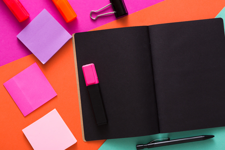 Stationery at modern creative work space. Stylish open notepad with black pages, paperclip, markers and memo notes on abstract multicolored background, top view, copy spaceの写真素材