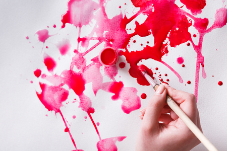 Artist creating watercolor painting. Brush and paint jar in female hands. Bright red drops on paper texture. Modern art backgroung with copy spaceの写真素材