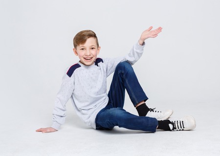 Portrait of happy smiling boy in braces sitting on the floor at white studio background. Kid in casual clothes posing on camera, copy spaceの写真素材