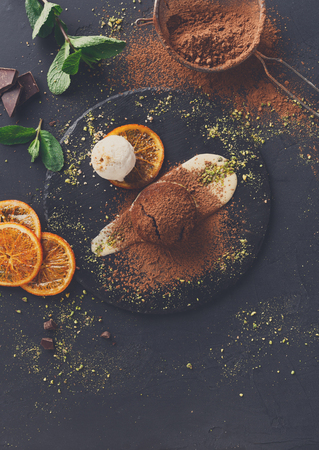 Exquisite french dessert. Chocolate fondant with creme anglaise and vanilla ice cream on round slate with pistachio crumble. Black background with cocoa, mint and sieve, top view, copy spaceの写真素材