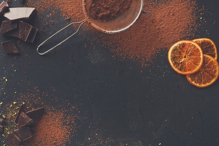Sweet background. Cocoa powder in a sieve and sprinkled on surface, orange citrons and chocolate pieces on black slate. Top view, copy spaceの写真素材