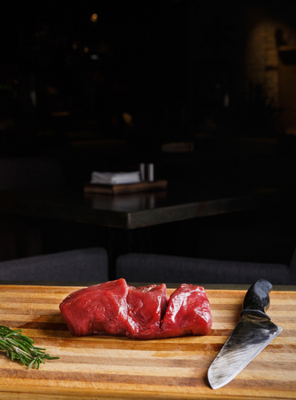 Filet mignon. Slices of fresh beef meat, rosemary and sharp knife on wooden cutting board at modern restaurant background, copy spaceの写真素材