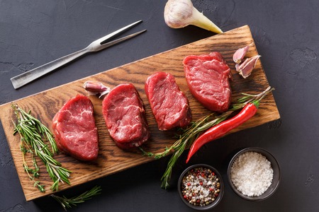 Raw filet mignon steaks with herbs and spices. Modern restaurant cuisine still life with fresh beef, salt, rosemary, garlic, chilli, meat fork on wooden board at black background, copy space, top viewの写真素材