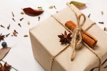 Holiday winter background. Gift box in craft paper decorated with cinnamon and anise star. Festive cold season composition of spices and dry leaves on white wood. Closeup, selective focus, copy spaceの写真素材