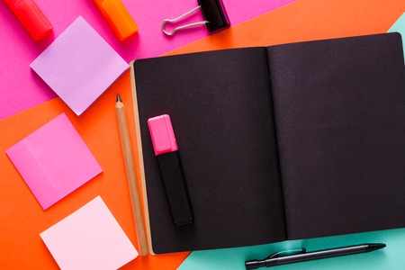 Stationery at modern creative work space. Stylish open notepad with black pages, paperclip, markers, pencil and memo notes on abstract multicolored background, top view, copy spaceの写真素材