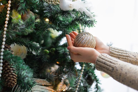 Female hands decorating christmas tree, holding golden bauble. Stylish modern design, balls, white beads and shiny garland. Winter holidays background with copy spaceの写真素材