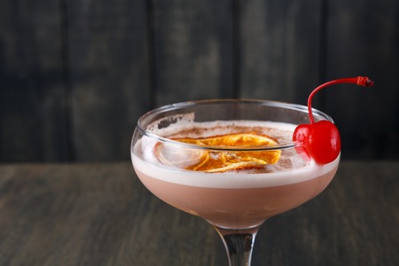 Modern bar cocktail at wooden background. Irish cream liquor in champagne glass decorated with maraschino cherry and orange slice closeup, copy spaceの写真素材