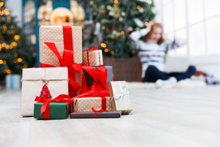 Winter holidays background. Christmas gift boxes band with red ribbons closeup at decorated xmas tree and happy young girl backdrop. Selective focus, copy spaceの写真素材