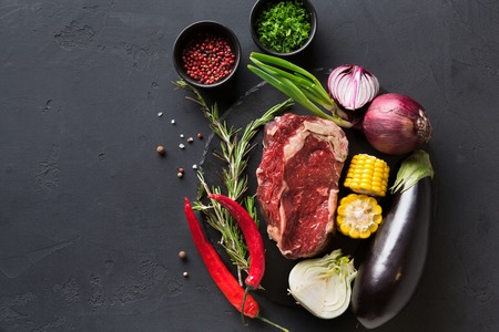 Raw rib eye steak with herbs and vegetables. Cooking ingredients for restaurant dish. Fresh meat, spices, eggplant, onion, chilli, corn on plate on black background, top view, copy spaceの写真素材