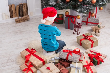Cute happy boy in santa hat unwrap christmas present box on holiday morning in beautiful room interior. Male child sitting with his back to camera near fir tree and fireplace. Winter holidays conceptの写真素材