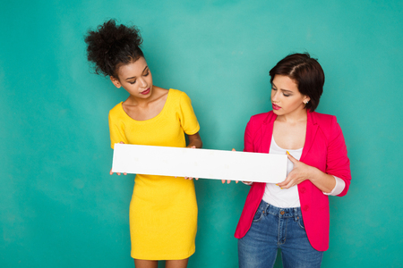 Two girlfriends in colorful clothes at blue background. African-american and caucasian women holding blank white board for copy spaceの写真素材