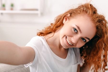 Pretty smiling girl making selfie, redhead in white clothes, closeupの写真素材
