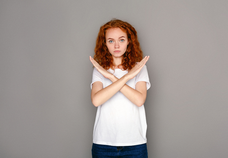 Redhead woman making stop sign with crossed hands. Serious girl denying, refusing any action, looking at camera, gray studio backgroundの写真素材