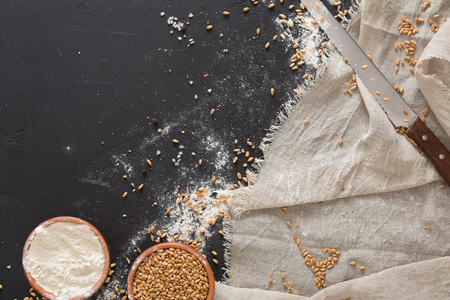 Baking background. Grain and flour in clay bowls, bread knife on linen cloth at black. Mockup for bread recepie or baking classes, top view, copy spaceの写真素材