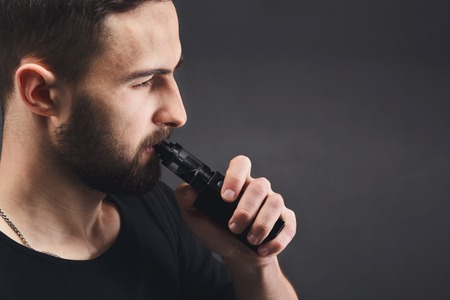 Man with vape at black studio background closeup. Young bearded guy smoking e-cigarette to quit tobacco. Vapor and alternative nicotine free smoking concept, copy space, closeupの写真素材