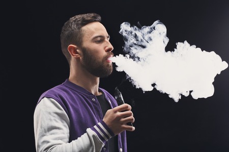 Man with vaping mod exhaling steam at black studio background. Bearded guy smoking e-cigarette to quit tobacco. Vapor and alternative nicotine free smoking concept, copy spaceの写真素材