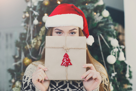 Young happy girl in red santa hat covering half face with christmas gift on xmas tree background. Funny and joyful winter holiday wallpaperの写真素材