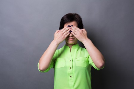 See no evil concept. Portrait of young scared woman covering eyes with hands, gray studio background.の写真素材