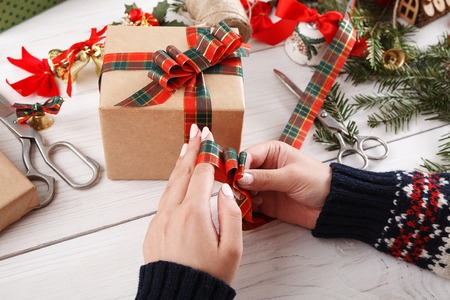 Creative diy hobby. Making bow on modern handmade xmas present, box in craft paper with red green checkered ribbon. Womans hands on white wood table with fir tree branches, gift decoration.の写真素材