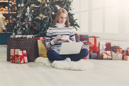 Christmas online shopping. Woman has coffee, buy presents on laptop, prepare to xmas, among gift boxes and packages. Winter salesの写真素材