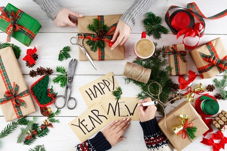 Creative diy hobby. Making modern handmade christmas present, boxes in craft paper, satin ribbon. Top view of two women's hands on messy white wood table with fir tree branches, bells, gift decorationの写真素材