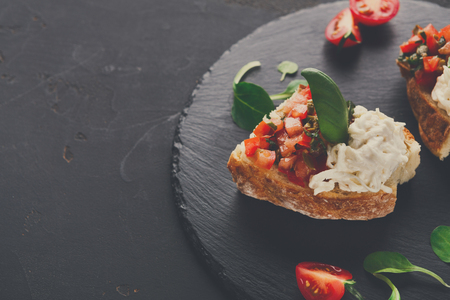Italian appetizer on black slate background. Crusty bruschetta with concasse tomatoes, stracciatella cheese decorated with spinach. Delicious and healthy restaurant meals, copy spaceの写真素材
