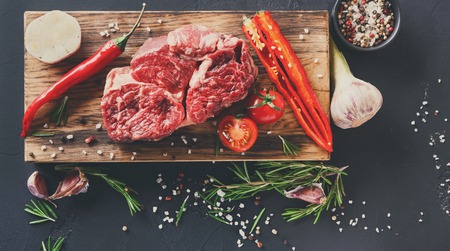 Raw rib eye steak with herbs and spices. Cooking ingredients for restaurant dish. Fresh meat, pepper salt, rosemary, chilli, cherry tomato and garlic on wooden board at black background, top viewの写真素材