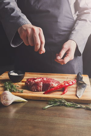 Man sprinkles filet mignon steaks with pepper salt. Chef working at open restaurant kitchen. Fresh meat, garlic and rosemary on wooden board. Modern restaurant cuisine backgroungの写真素材