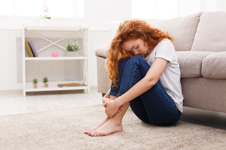 Young lonely redhead woman sitting on the floor at home. Thoughtful, sad woman in casual clothes hiding from the worldの写真素材
