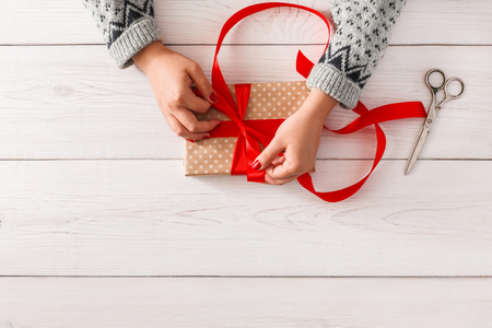 DIY hobby. Womans hands wrapping christmas or other holiday handmade present in paper with red ribbon. Making bow at present box, decoration of gift. Scissors on white wooden table, top view.の写真素材