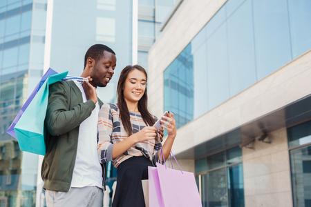 Multiethnic happy couple reading message in smartphone after shopping together, walking with bags at mall, copy spaceの写真素材