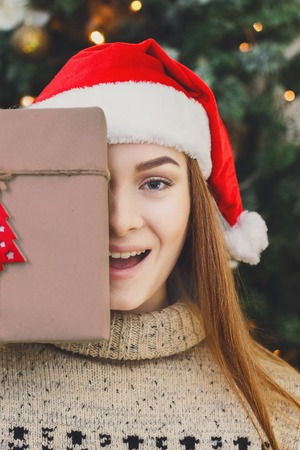 Young happy girl in red santa hat covering half face with christmas gift on xmas tree background. Funny and joyful winter holiday wallpaperの写真素材