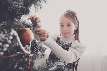 Pretty little girl decorate big christmas tree. Child prepare to winter holiday, hang decorations, tinsel, bead garland, ornaments, baubles and balls. Christmas atmosphere at cosy homeの写真素材