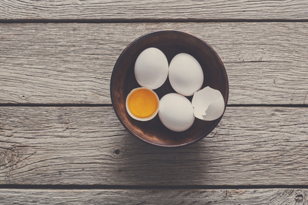 Fresh chicken white home eggs with cracked eggshell and yolk in bowl at rustic wood table. Top view...