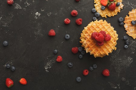 Belgium waffles with berries on black background sprinkled with flour. Border of checkered cookies top view with copy space. Cooking sweet food and tasty breakfast conceptの写真素材