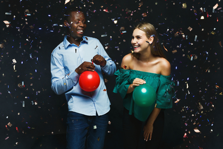 Multiethnic couple having fun. Young man and woman with balloons at black background, showered with confetti. Birthday or new year party celebration conceptの写真素材